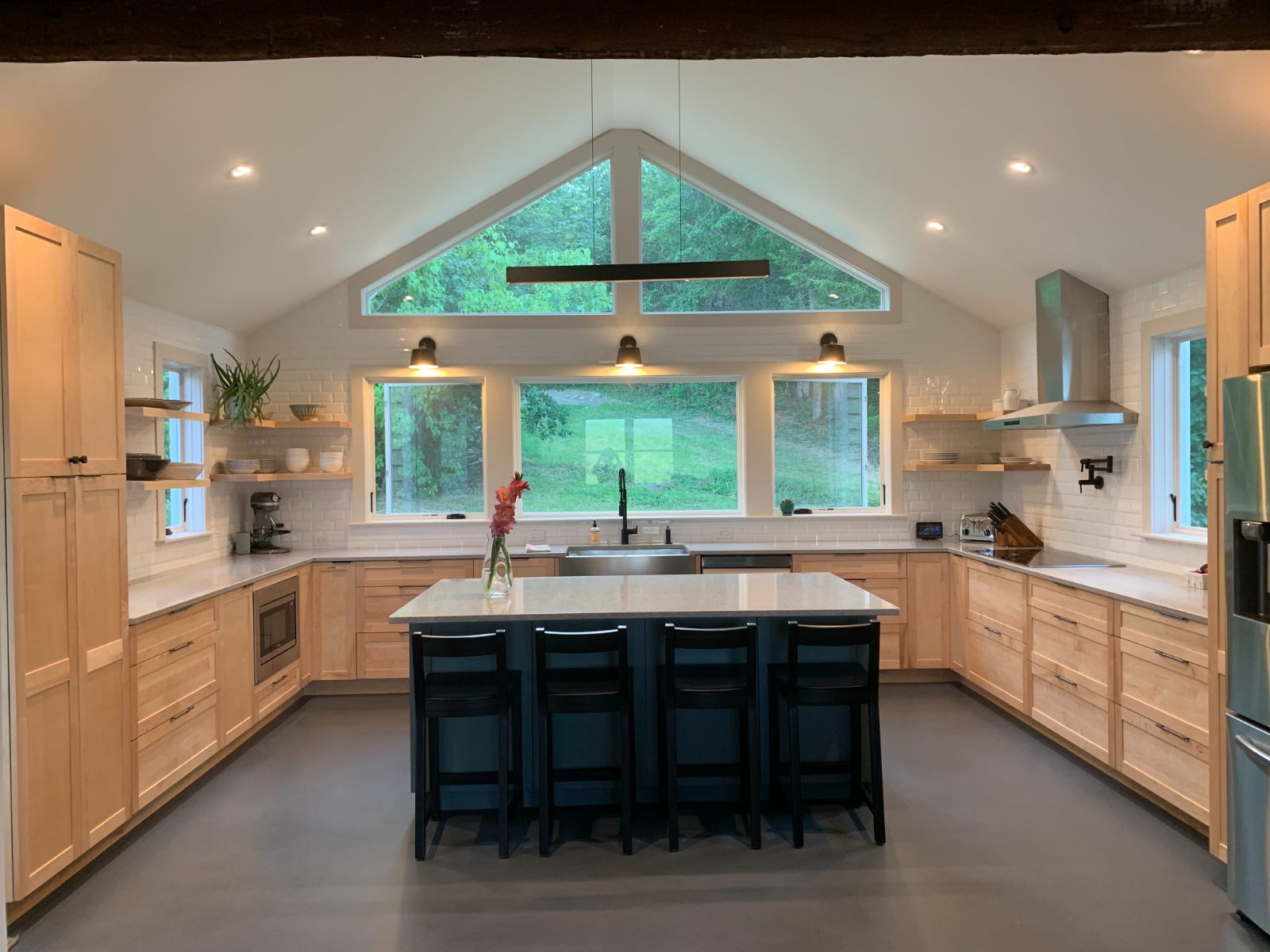 Natural maple cabinetry with slate-blue island and black stools under vaulted windows